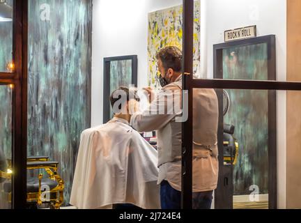 Un barbiere in maschera facciale che lavora su un taglio di capelli in barbiere maschile a Siviglia, Andalusia, Spagna. Sevillan spagnolo parrucchieri fare capelli taglio. Foto Stock