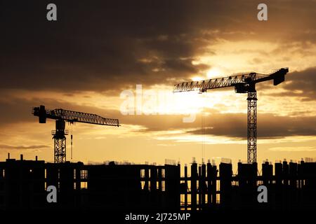 Sagome di gru a torre e lavoratori su ponteggi di edificio incompiuto all'alba. Edilizia abitativa, condominio in città sullo sfondo Foto Stock
