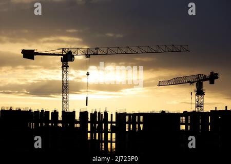 Sagome di gru a torre e lavoratori su ponteggi di edificio incompiuto all'alba. Edilizia abitativa, condominio in città sullo sfondo Foto Stock