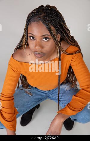 Studio shot di una giovane donna nera con vintage top arancione e i jeans larghi in posa su uno sfondo bianco squatting Foto Stock