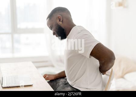 Mal di schiena afroamericano ragazzo con mal di schiena che massaggiano il mal di schiena più basso, che soffre di dolore mentre si siede a casa sul posto di lavoro. Dolore lombare. Prob. Salute Foto Stock