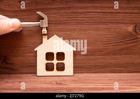 Dita che tengono un martello in acciaio miniaturizzato su un piccolo modello di casa piatta su uno sfondo di legno. Home progetto di revamping. Foto Stock