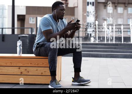 Giovane atletico africano in abbigliamento sportivo riposato dopo l'allenamento in centro, seduto su panca, SMS su smartphone e ascoltare musica Foto Stock