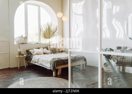 Interno di camera da letto moderna e soleggiata con divisorio in plastica trasparente. Letto in legno rivestito con biancheria da letto grigia, tela di cotone beige con frangia sul bro Foto Stock