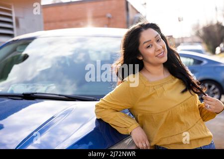 Giovane donna ispanica sicura appoggiata sulla sua auto all'esterno, con camicia gialla. Foto Stock