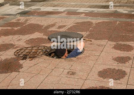 Un lavoratore si arrampica fuori da un tombino aperto nella strada. Pericoloso botola aperta non protetta sulla strada. Incidente con una botola fognaria in città. Il con Foto Stock