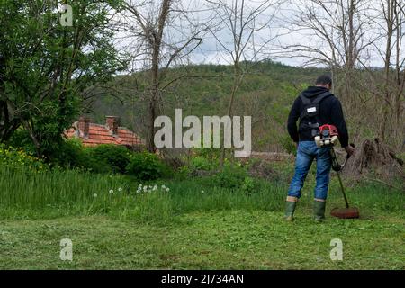 Un uomo falcia l'erba con un trimmer in una giornata intensa. Coltivatore falciando erba in un villaggio nel cortile con mano prato a benzina tosaerba Foto Stock