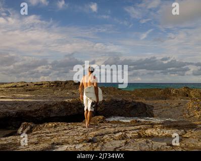 Vista posteriore del surfista che trasporta la tavola da surf che cammina attraverso la superficie rocciosa verso l'oceano Foto Stock