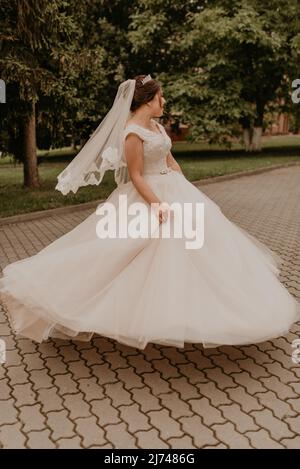 Giovane donna caucasica europea dai capelli scuri sposata in abito da sposa bianco con velo lungo e tiara sulla testa che girano vortici in posa a ballare Foto Stock