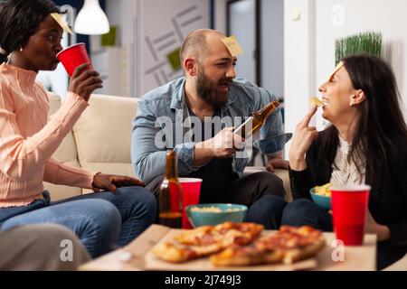 Un team diversificato di compagni di lavoro che si divertono con i charades e le note appiccicose al party dopo il lavoro. L'uomo e le donne che giocano indovinare che giocano in ufficio festeggiamenti dopo le ore, bevendo birra. Foto Stock