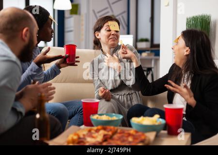 Gruppo di colleghi diversi che giocano a Guess Who con note appiccicose sulla fronte, godendo di attività divertente con gioco di carades. La gente indovina pantomima alla festa delle bevande dopo l'orario di lavoro. Foto Stock