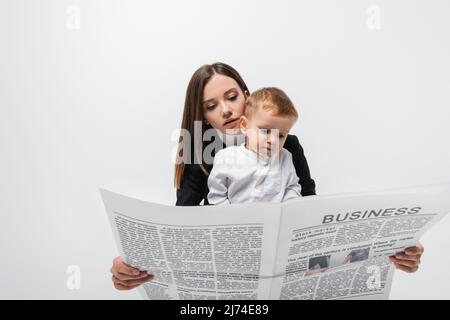 giovane donna d'affari che legge il giornale vicino bambino piccolo isolato su grigio Foto Stock