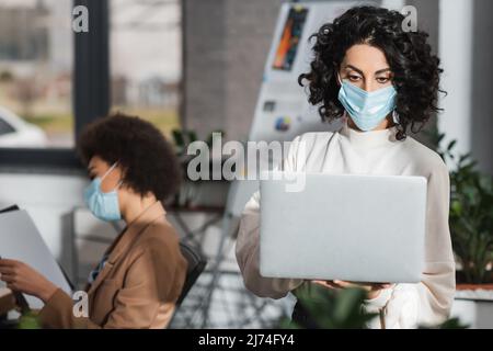 Donna d'affari musulmana in maschera medica usando il laptop vicino alla blurred african american collega in ufficio Foto Stock