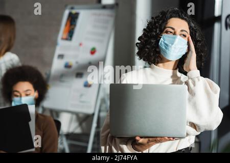 Donna d'affari musulmana in maschera medica che tiene il laptop mentre soffre di mal di testa in ufficio Foto Stock