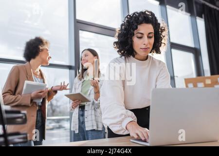 Donna d'affari musulmana che usa il laptop vicino ai colleghi multietnici offuscati che parlano in ufficio Foto Stock