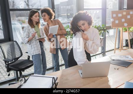 Donna d'affari musulmana che parla sullo smartphone e utilizza un laptop vicino a colleghi interracial offuscati in ufficio Foto Stock