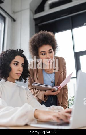 Donna d'affari afroamericana che tiene la cartella della carta vicino al collega arabo usando il laptop Foto Stock