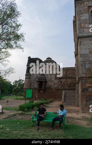 Due giovani uomini seduti su una panchina verde accanto ad edifici in pietra erosi in un complesso di patrimonio storico in India Foto Stock