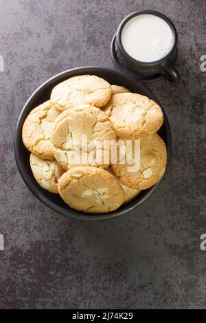 Biscotti Macadamia appena fatti con cioccolato bianco in primo piano nella ciotola nera e una tazza di latte sul tavolo da vicino. Vista dall'alto verticale Foto Stock