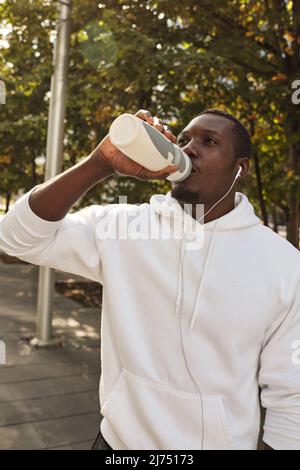 Giovane atletico uomo nero sete indossando la felpa bianca che beve l'acqua dalla bottiglia di sport dopo le lunghe distanze che corre all'aperto Foto Stock