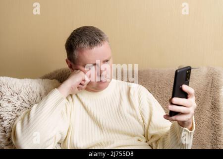 Un uomo guarda pensieroso in uno smartphone, poggiando la testa sulla mano. Europeo in un maglione bianco si siede su un divano con un telefono in mano. Foto Stock