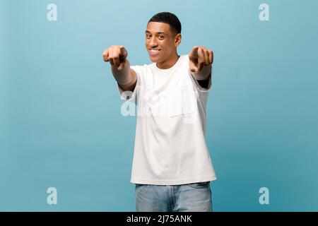 Ehi. Testa di allegro sereno spensierato giovane afro-americano in casual bianco t-shirt punti dita di entrambe le mani a voi, studio ritratto isolato su sfondo blu Foto Stock