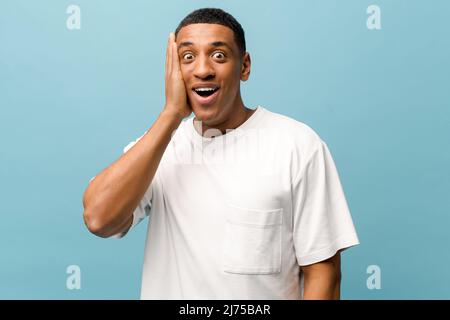 Stordito ragazzo afroamericano guardando la macchina fotografica con bocca aperta, sorpreso con incredibile prezzo di vendita multirazziale giovane ragazzo tenendo faccia con la palma, shock maschio isolato sul muro blu Foto Stock