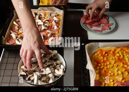 Defocalizzare gli ingredienti per la pizza sull'impasto. Cucina a mano femminile e maschile. Salsa di pomodoro rosso, salsicce, mais dolce, funghi. Pizza italiana fatta in casa. Base Foto Stock