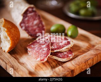 Salame affettato su un tagliere con pane; ciotola di olive verdi in background Foto Stock