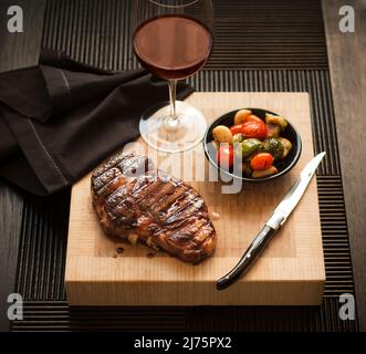 Bistecca intera su un tagliere con una ciotola di verdure arrosto e un bicchiere di vino rosso Foto Stock