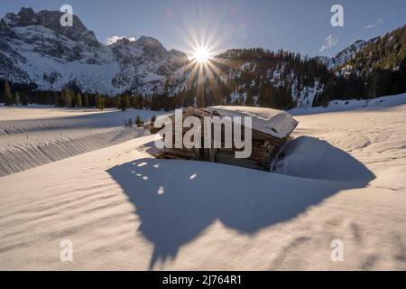 Una piccola capanna in legno nella Valle del Rodano, vicino a Hinterriss nel Karwendel, una parte delle Alpi in Austria in inverno con neve. Il sole è basso sulle montagne con le Karwendelspitze orientali, sotto un cielo prevalentemente blu e il tempo giusto, la capanna getta la sua ombra verso lo spettatore, sullo sfondo il paesaggio nella neve. Foto Stock