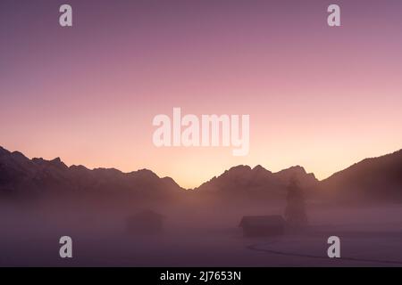 Atmosfera nebbia tra Krün e Wallgau in Werdenfelser Land in inverno. La fitta nebbia si trova sui prati nevosi con Stadln, sullo sfondo le montagne di Wetterstein e le Zugspitze in viola sera rosso con cielo senza nuvole. Foto Stock