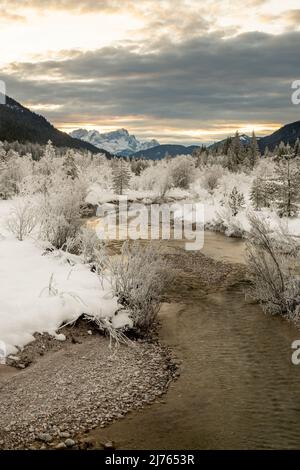 Un piccolo torrente laterale dell'Isar, si snoda attraverso il paesaggio innevato coperto di gelo in inverno. Sullo sfondo le Zugspitze e la luce soffusa della sera. Foto Stock