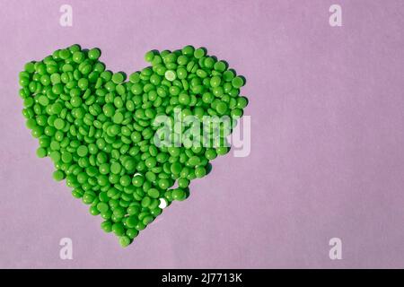 Splendidi granuli di cera verde con estratto di aloe per la depilazione vengono versati su sfondo viola chiaro, a forma di cuore. Epilazione, depilazione, Foto Stock