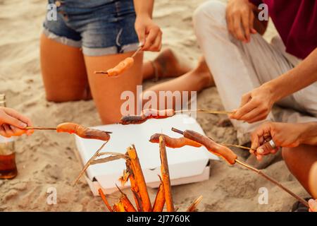 gara mista gruppo multinazionale di amici che cucinano salsicce e bevono limonata al tramonto sulla sabbia Foto Stock