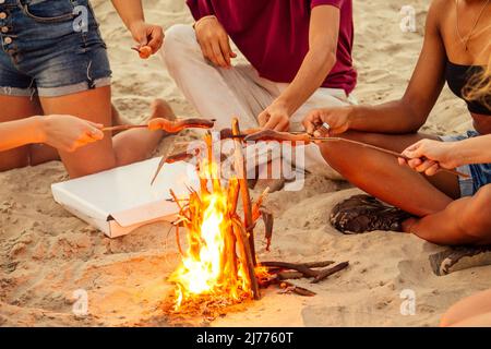 gara mista gruppo multinazionale di amici che cucinano salsicce e bevono limonata al tramonto sulla sabbia Foto Stock