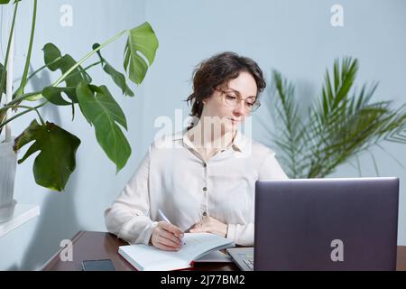 Una donna d'affari con gli occhiali scrive in un taccuino al suo posto di lavoro Foto Stock