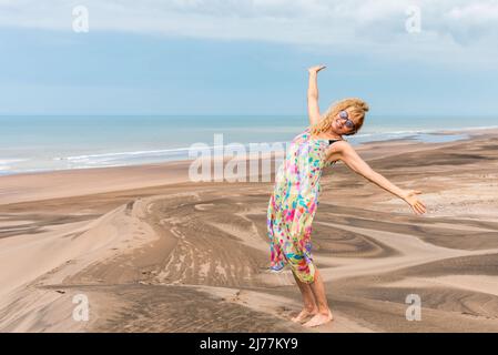 Donna in abito guardando la macchina fotografica mentre alza le braccia gesturando la felicità da una duna del deserto Foto Stock