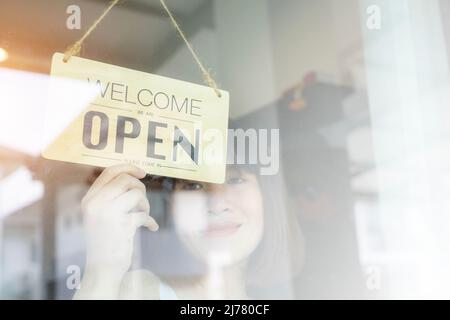 Donna asiatica proprietario negozio girare aprire cartello attraverso il vetro della porta e pronto per il servizio. Foto Stock