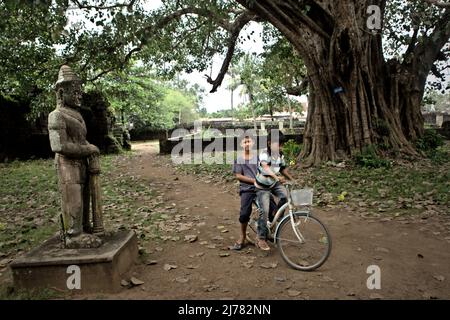 Bambini in bicicletta vicino a una scultura dvarapala a Banteay Prei Nokor, un antico tempio a Kampong Cham, Cambogia. Foto Stock