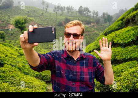 Bloger rosso zenzero maschio godendo mattina scattando le immagini selfie sulla macchina fotografica di smartphone in India piantagioni di chai Munnar Foto Stock