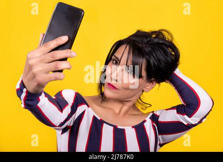 Bella donna indossa una t-shirt a righe. Giovane donna bruna sta prendendo un selfie con il suo telefono cellulare su uno sfondo giallo. Foto Stock