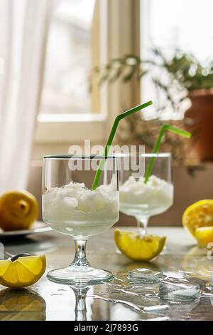 Sorbetto al limone in due bicchieri con limoni, cubetti di ghiaccio e cannucce da bere su sfondo grigio e lato posteriore della finestra Foto Stock