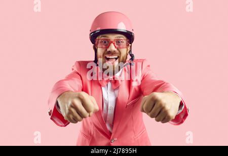 L'uomo divertente in casco protettivo rosa finge di essere motociclista isolato su sfondo rosa. Foto Stock