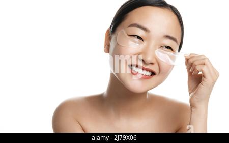 Cosmetici per la cura della pelle. Donna asiatica sorridente applica sotto gli occhi anti-puffiness patch per una pelle sana e fresca del viso, in piedi su sfondo bianco Foto Stock