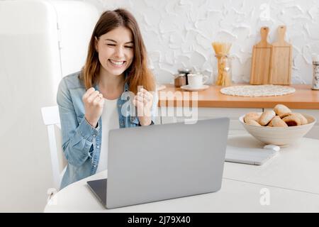 Gioiosa giovane donna d'affari è felice del suo successo di lavoro, sorridendo guardando il laptop. Bella freelancer femminile o studente ha ottenuto una promozione di lavoro Foto Stock