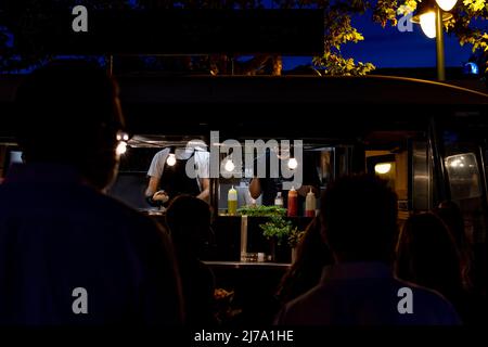 2 cuochi irriconoscibili in un furgone illuminato di notte, preparando fast food per diversi clienti irriconoscibili con le spalle girate in fila Foto Stock