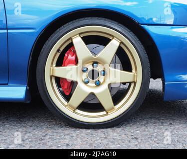 Primo piano di un cerchio in lega d'oro metallica a 6 razze, pinze dei freni rosse a 4 pistoni e dischi dei freni scanalati. Auto blu metallizzato su strada asfaltata. Nessun logo. Foto Stock