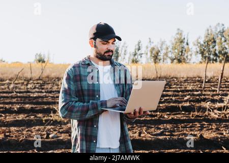 Giovane contadino latino telelavoro sul suo portatile nel mezzo della sua terra. Sostenibilità agricola Foto Stock