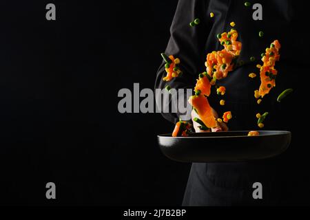 Uno chef professionista prepara pezzi di pesce rosso con verdure in una padella. Cibo in volo congelato su sfondo nero. Ricette per cucinare fi Foto Stock